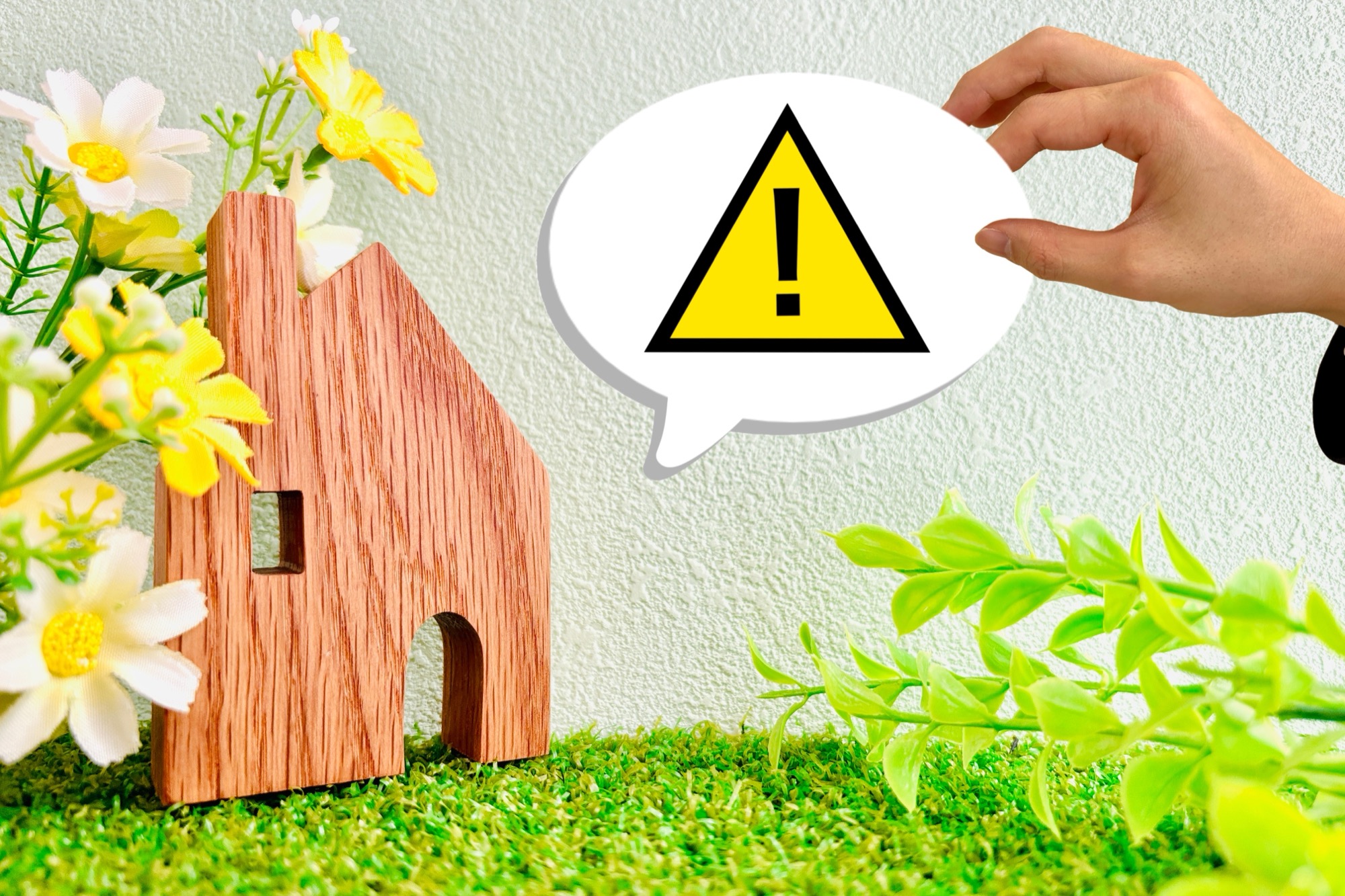 緑の中に置かれた住宅の形をした木の板と、手が添えられた注意マークの吹き出し