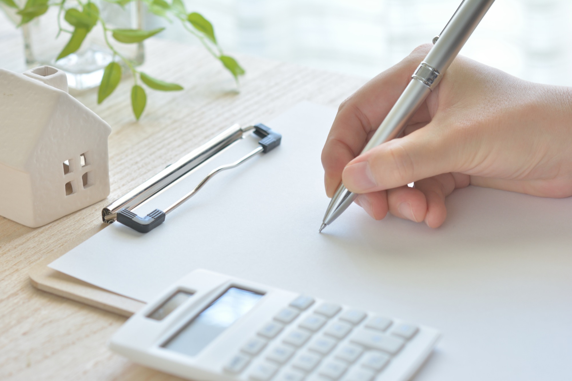白紙にメモする女性の手元、電卓と家の模型